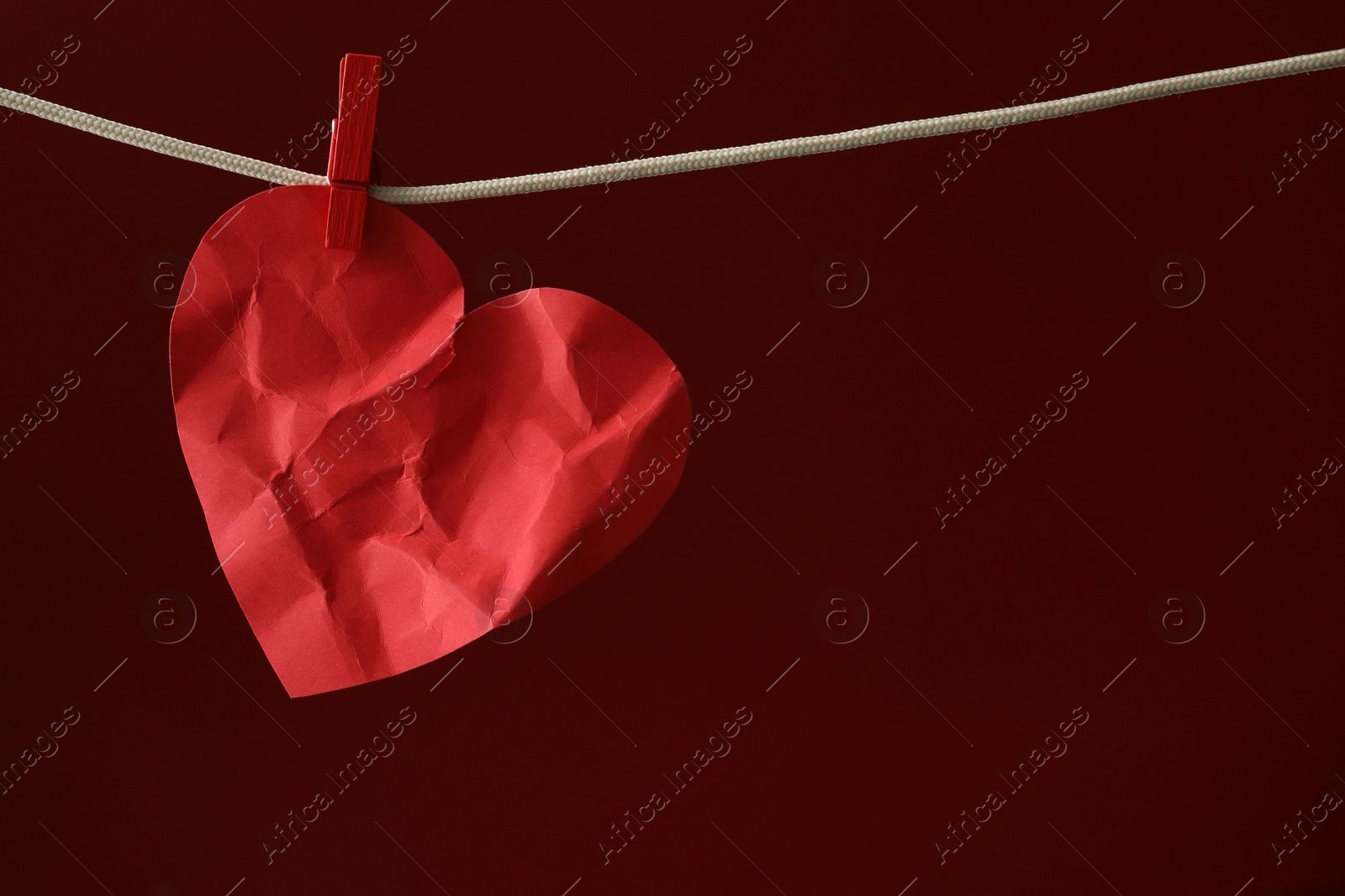 Photo of Crumpled red paper heart on rope against burgundy background, space for text. Broken heart