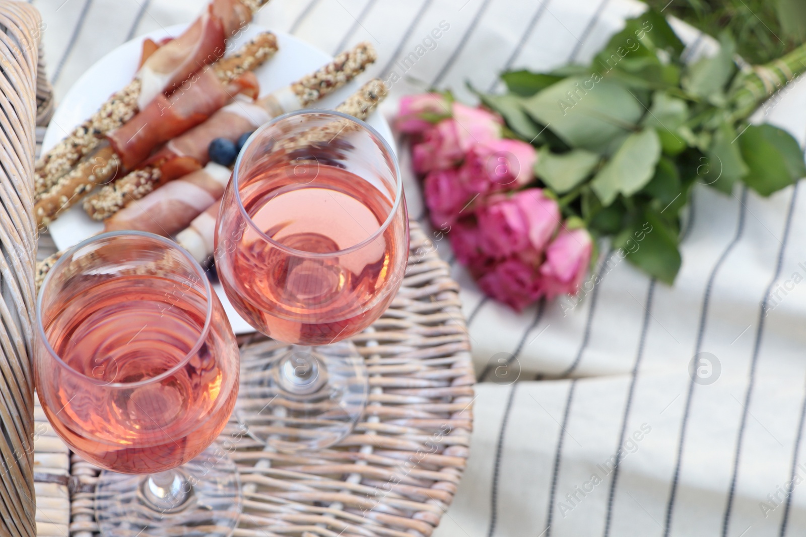 Photo of Glasses of delicious rose wine, food, flowers and basket on picnic blanket outdoors, closeup