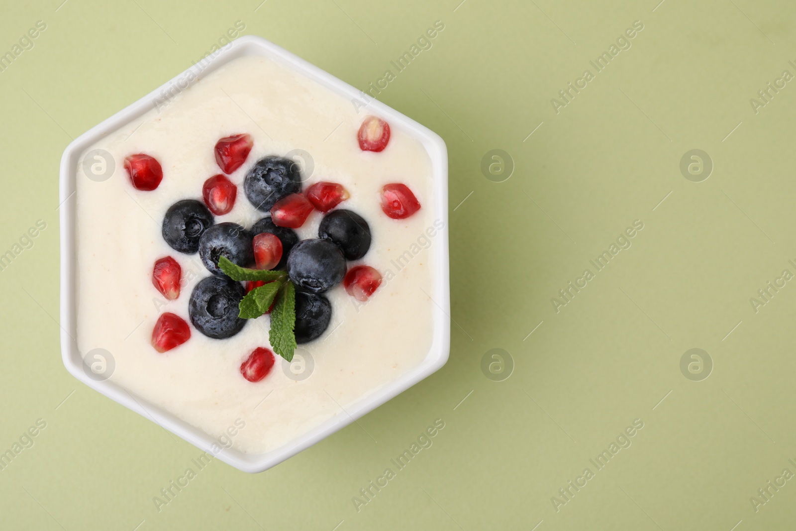 Photo of Bowl of delicious semolina pudding with blueberries, pomegranate and mint on light green background, top view. Space for text