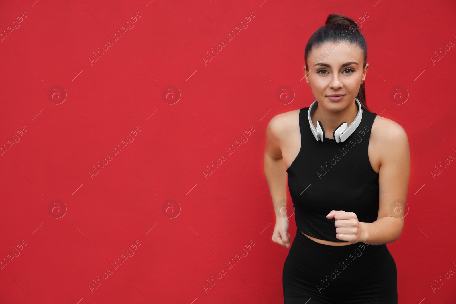 Photo of Young woman in sportswear with headphones running on red background. Space for text