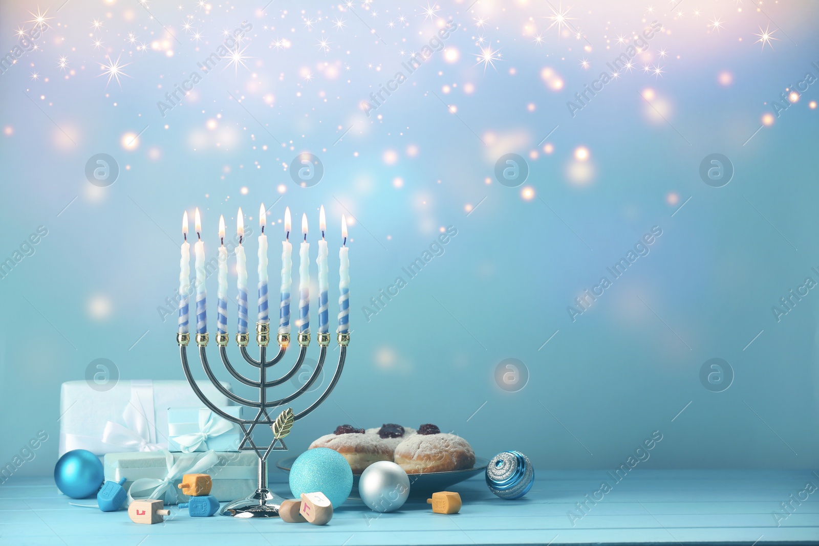 Image of Hanukkah celebration. Menorah with burning candles, dreidels, donuts and gift boxes on light blue wooden table, space for text
