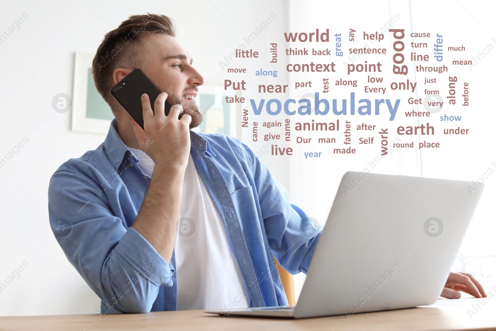 Image of Young man talking on mobile phone while working with laptop at desk, word cloud in front of him