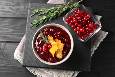 Photo of Fresh cranberry sauce, rosemary, orange peel and berries on black wooden table, top view