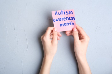 Photo of Woman holding sheet of paper with words AUTISM AWARENESS MONTH on grey background, top view