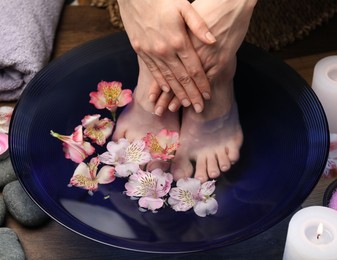 Photo of Woman soaking her feet in bowl with water and flowers on floor, closeup. Spa treatment