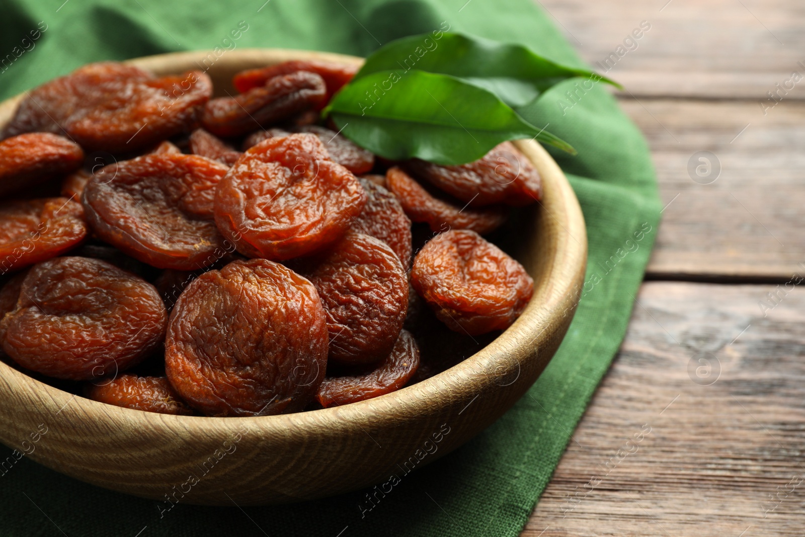 Photo of Bowl of tasty apricots and green leaves on wooden table, closeup. Dried fruits