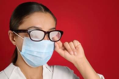 Photo of Woman wiping foggy glasses caused by wearing medical mask on red background, closeup