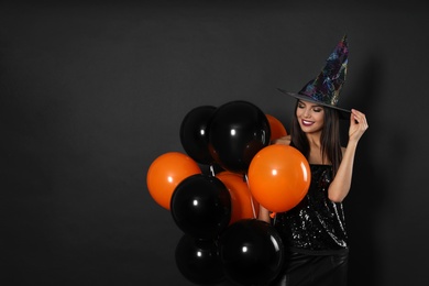 Beautiful woman wearing witch costume with balloons for Halloween party on black background, space for text