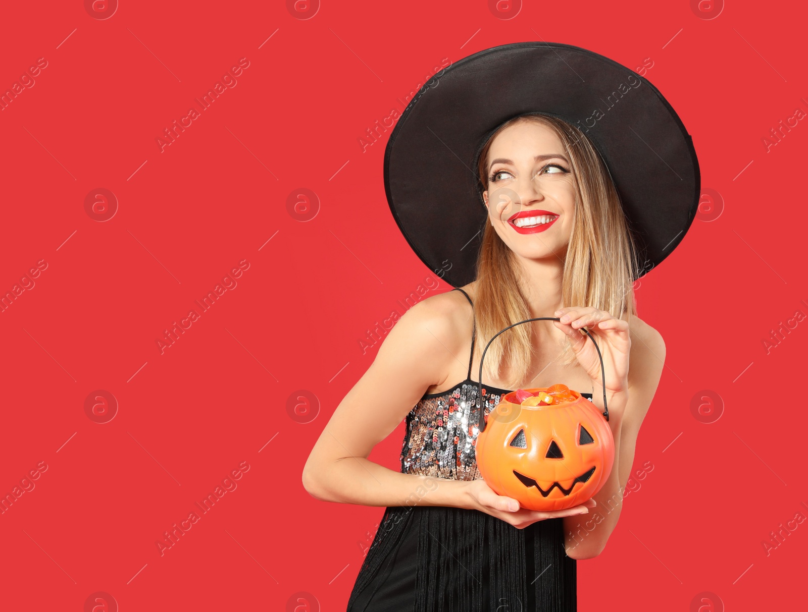 Photo of Beautiful woman wearing witch costume with Jack O'Lantern candy container on red background, space for text. Halloween party