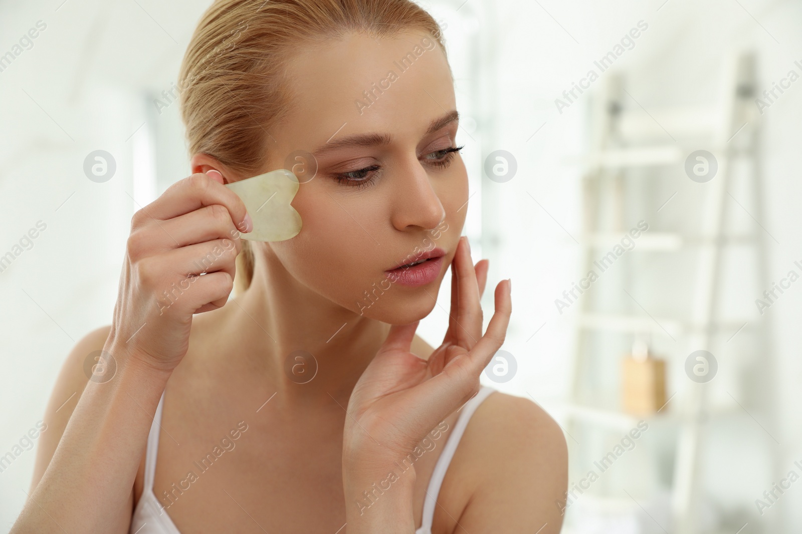 Photo of Beautiful young woman doing facial massage with gua sha tool in bathroom, closeup