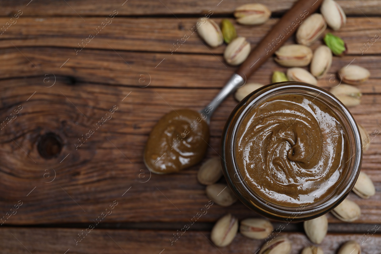 Photo of Tasty pistachio nut paste in jar and spoon on wooden table, flat lay. Space for text