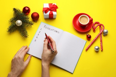 Photo of Woman filling list of New Year's resolutions in notebook on yellow background, top view