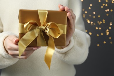 Photo of Christmas present. Woman holding gift box against grey background with blurred lights, closeup