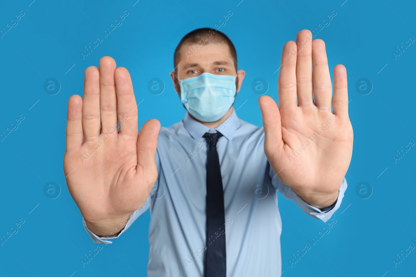 Photo of Man in protective mask showing stop gesture on light blue background. Prevent spreading of coronavirus