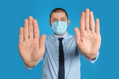 Photo of Man in protective mask showing stop gesture on light blue background. Prevent spreading of coronavirus