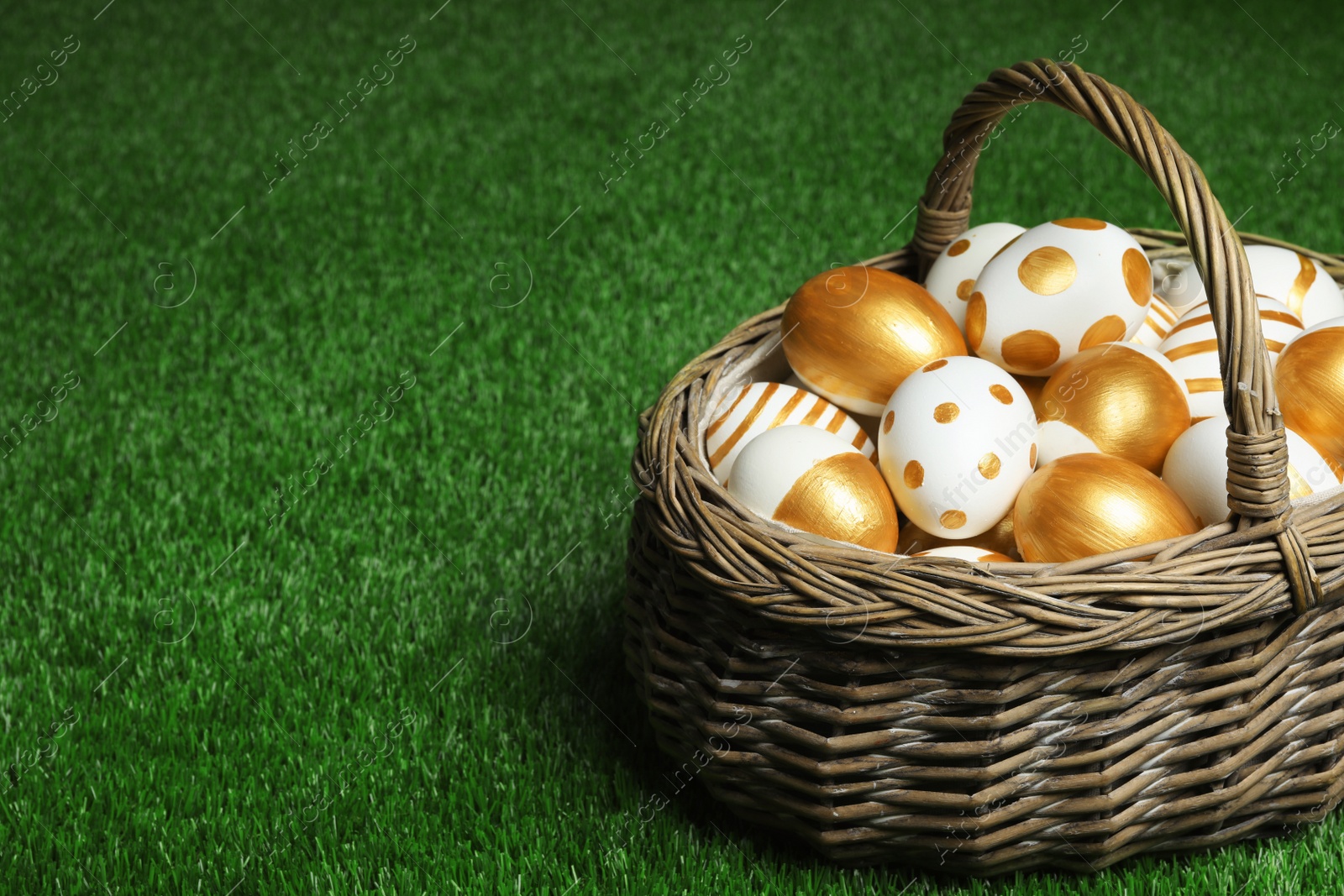 Photo of Wicker basket of traditional Easter eggs decorated with golden paint on green lawn, space for text