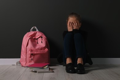 Photo of Upset girl with backpack sitting on floor near black wall. School bullying