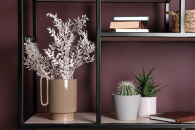 Photo of Stylish ceramic vase with dry plants and houseplants on shelf near brown wall