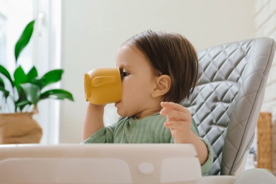 Cute little baby drinking from cup in high chair indoors