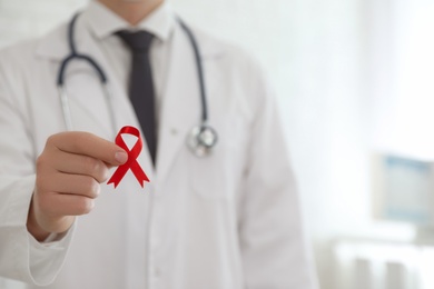 Doctor holding red ribbon on blurred background, closeup with space for text. AIDS awareness month