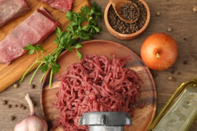 Meat grinder with beef, garlic, onion, parsley, oil and peppercorns on wooden table, flat lay