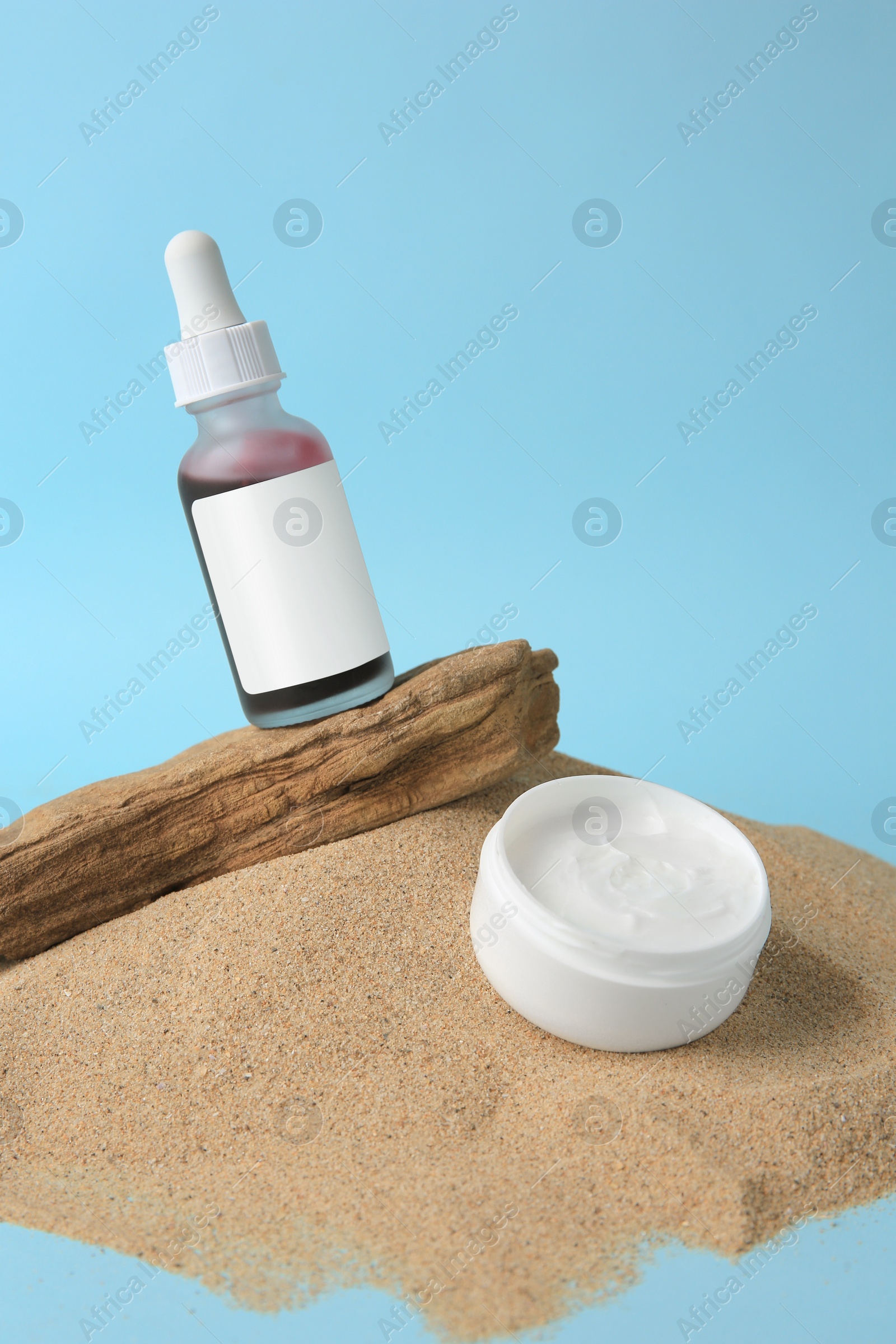 Photo of Bottle with serum and jar of cream on sand against light blue background. Cosmetic product