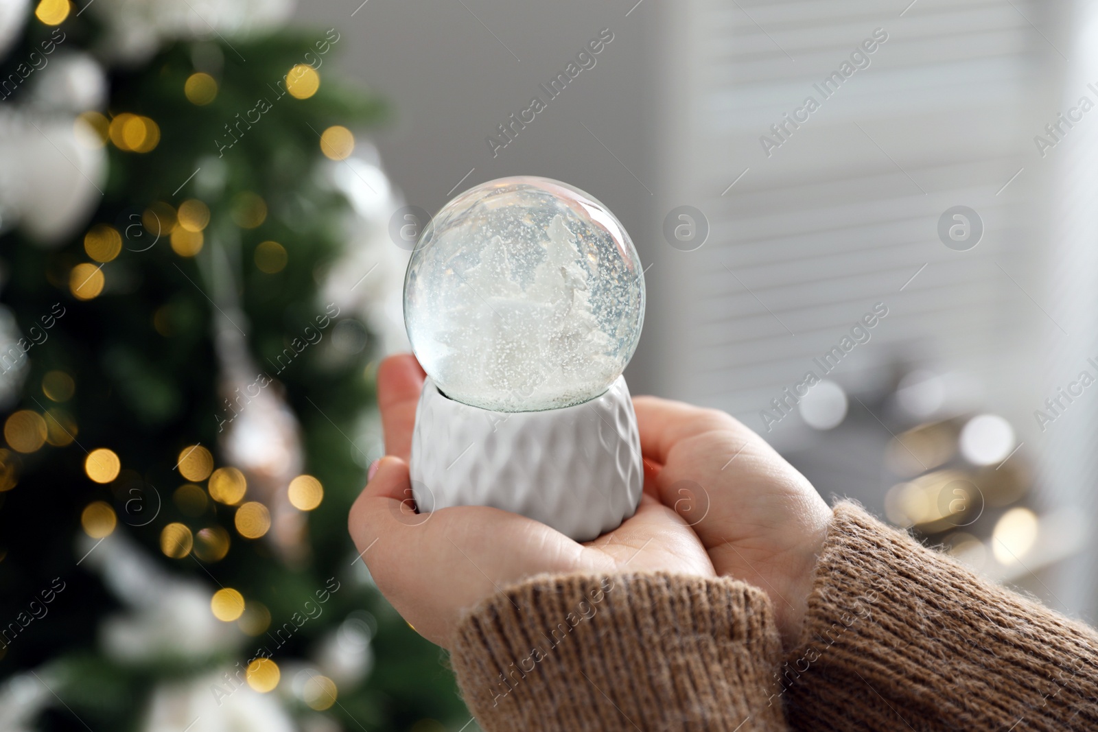 Photo of Woman in sweater holding decorative snow globe near Christmas tree, closeup