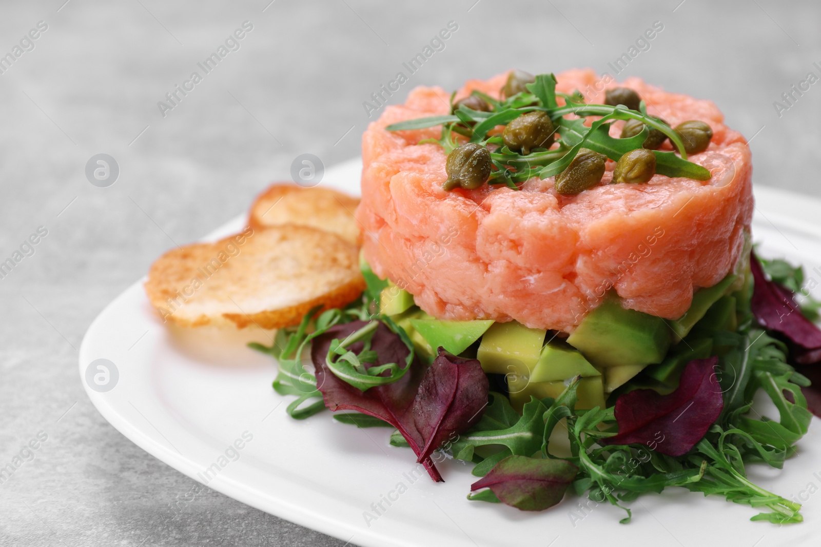 Photo of Tasty salmon tartare with avocado, greens and croutons on grey table, closeup