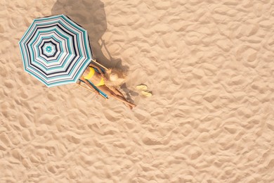 Woman resting in sunbed under striped beach umbrella at sandy coast, aerial view. Space for text