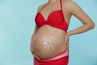 Young pregnant woman with sun protection cream on belly against light blue background, closeup