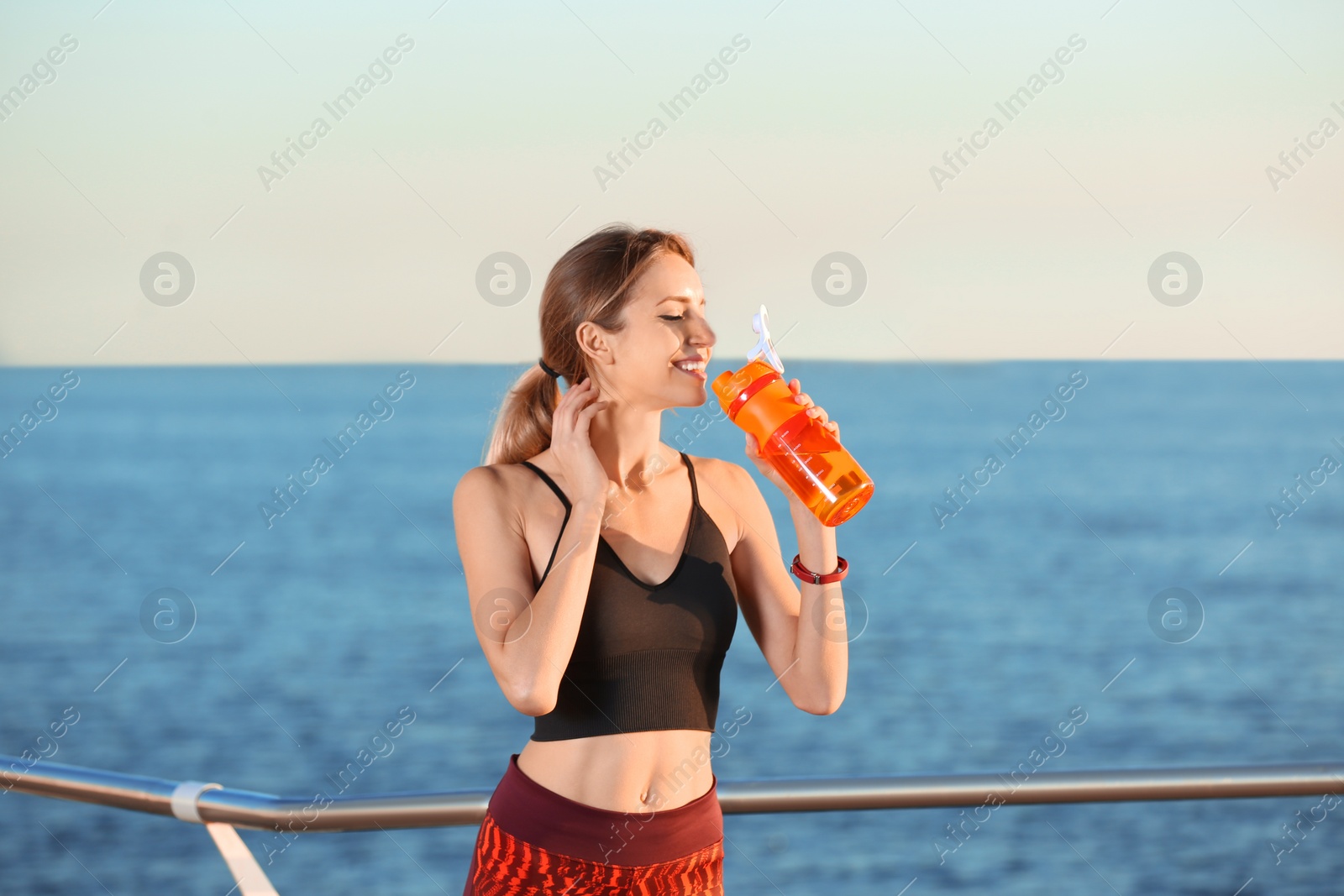 Photo of Young woman drinking water from bottle after fitness exercises on pier in morning