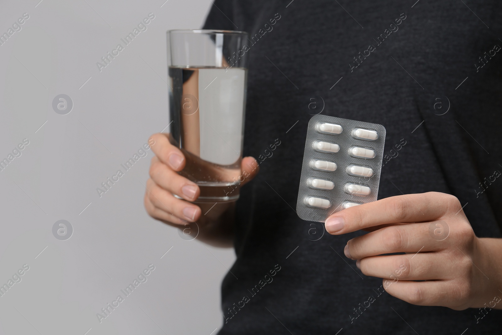 Photo of Woman holding pills in blister pack and glass of water on gray background, closeup. Space for text