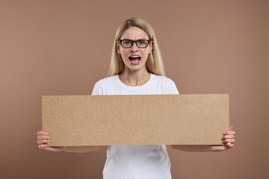 Angry woman holding blank cardboard banner on brown background, space for text
