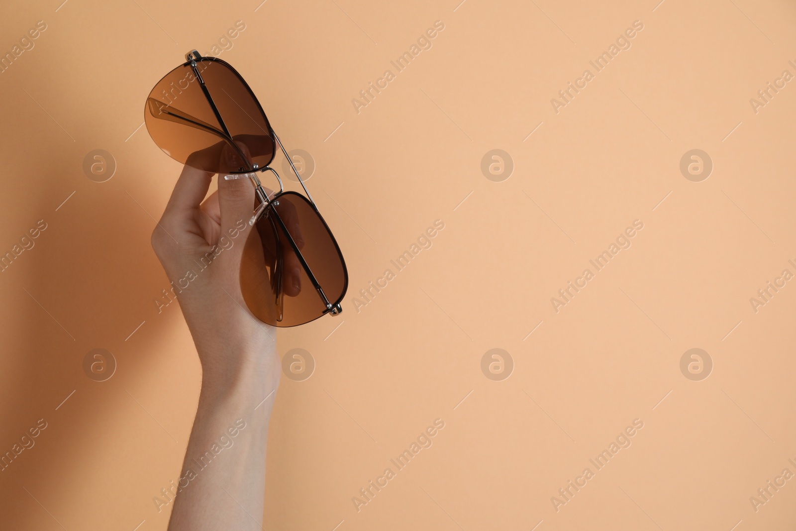 Photo of Woman holding stylish sunglasses on pale orange background, closeup. Space for text