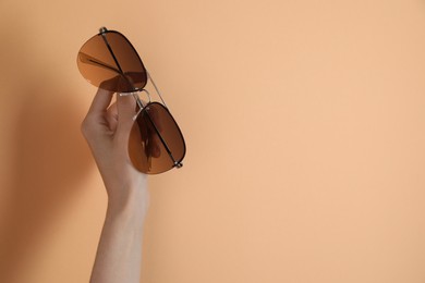 Photo of Woman holding stylish sunglasses on pale orange background, closeup. Space for text