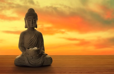 Image of Beautiful stone Buddha sculpture with flower petals on wooden surface at sunset. Space for text