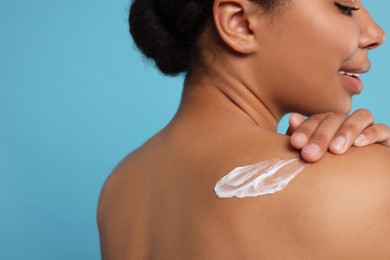 Young woman applying body cream onto back on light blue background, closeup