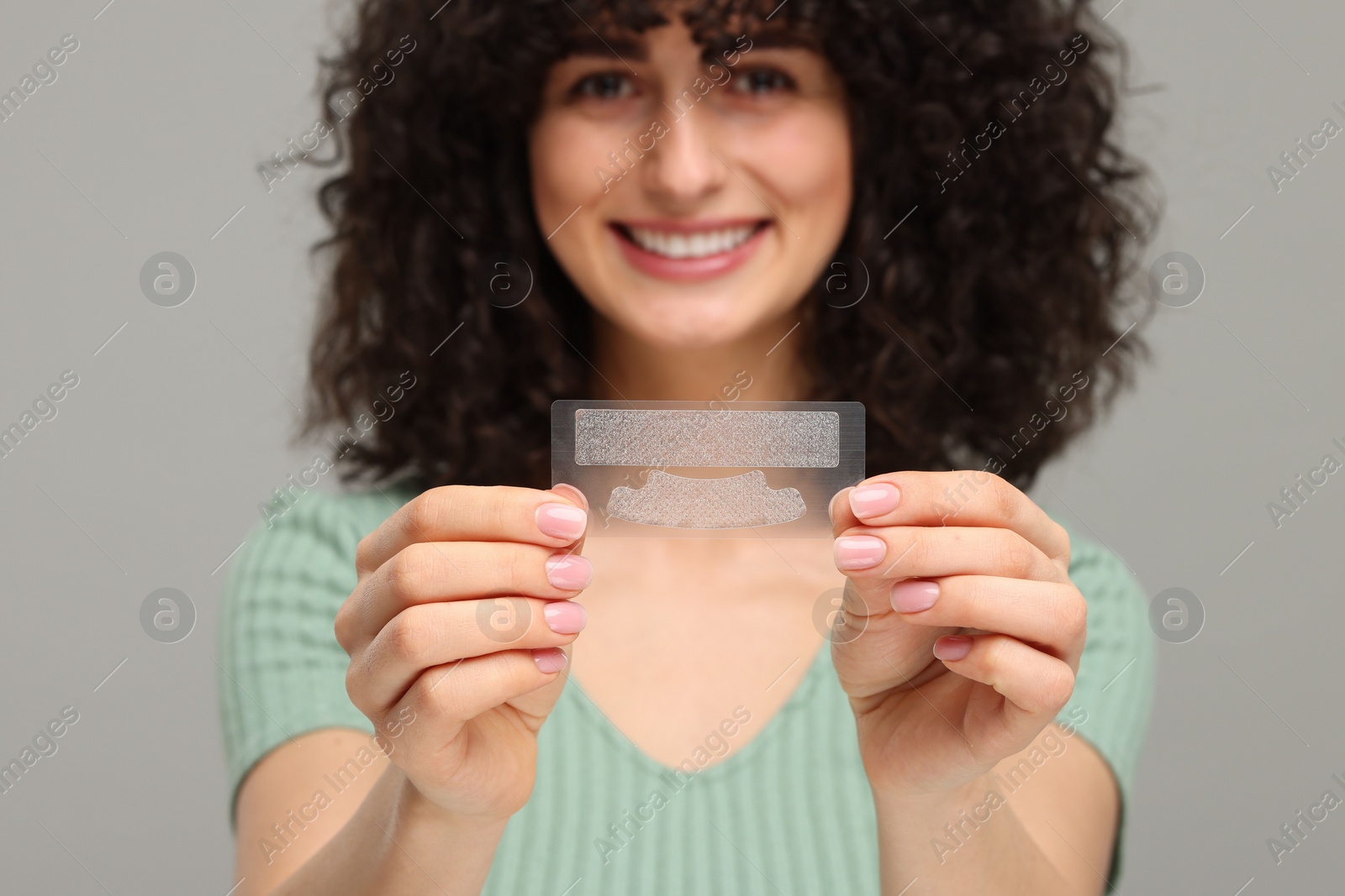 Photo of Young woman holding teeth whitening strips on grey background, selective focus