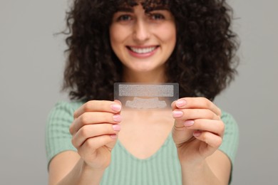 Young woman holding teeth whitening strips on grey background, selective focus