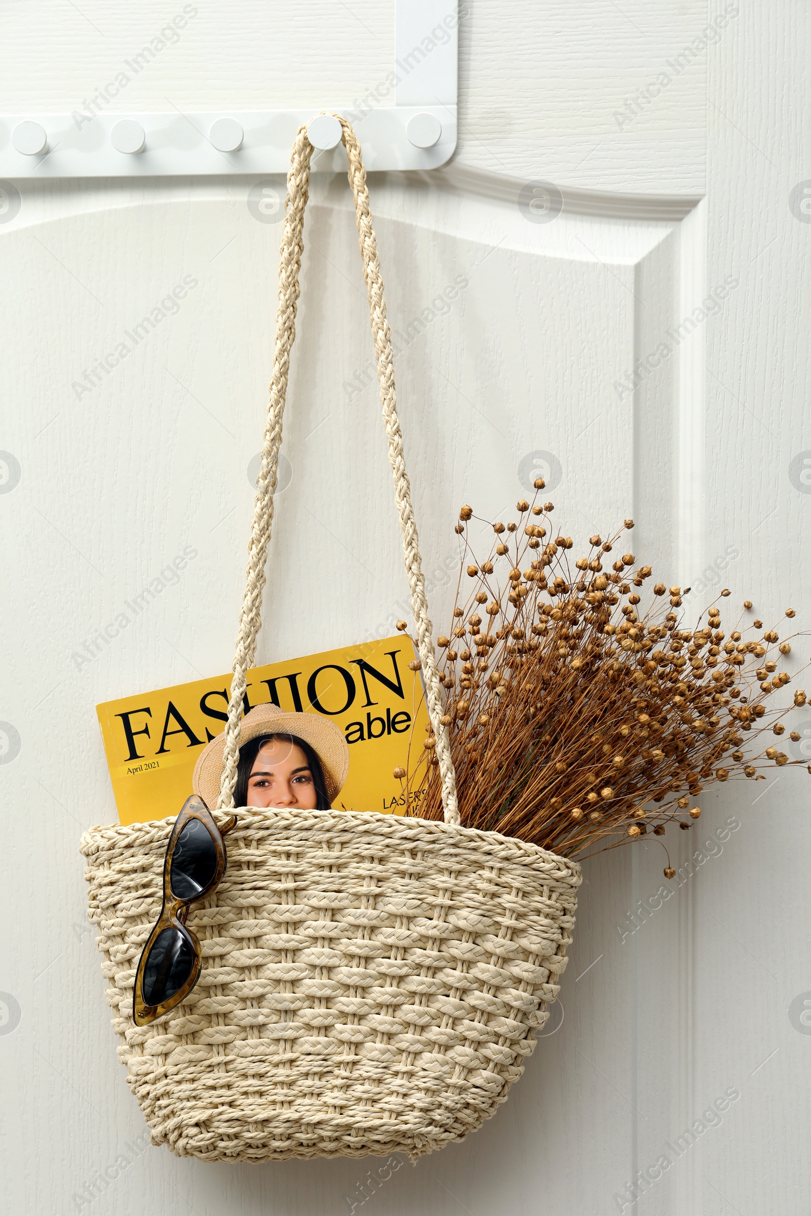 Photo of Stylish beach bag with beautiful dried flowers, sunglasses and magazine hanging on white wooden door