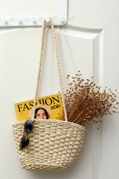 Stylish beach bag with beautiful dried flowers, sunglasses and magazine hanging on white wooden door