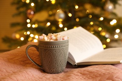 Photo of Cup of tasty cocoa with marshmallows and open book on pink fabric against blurred Christmas tree