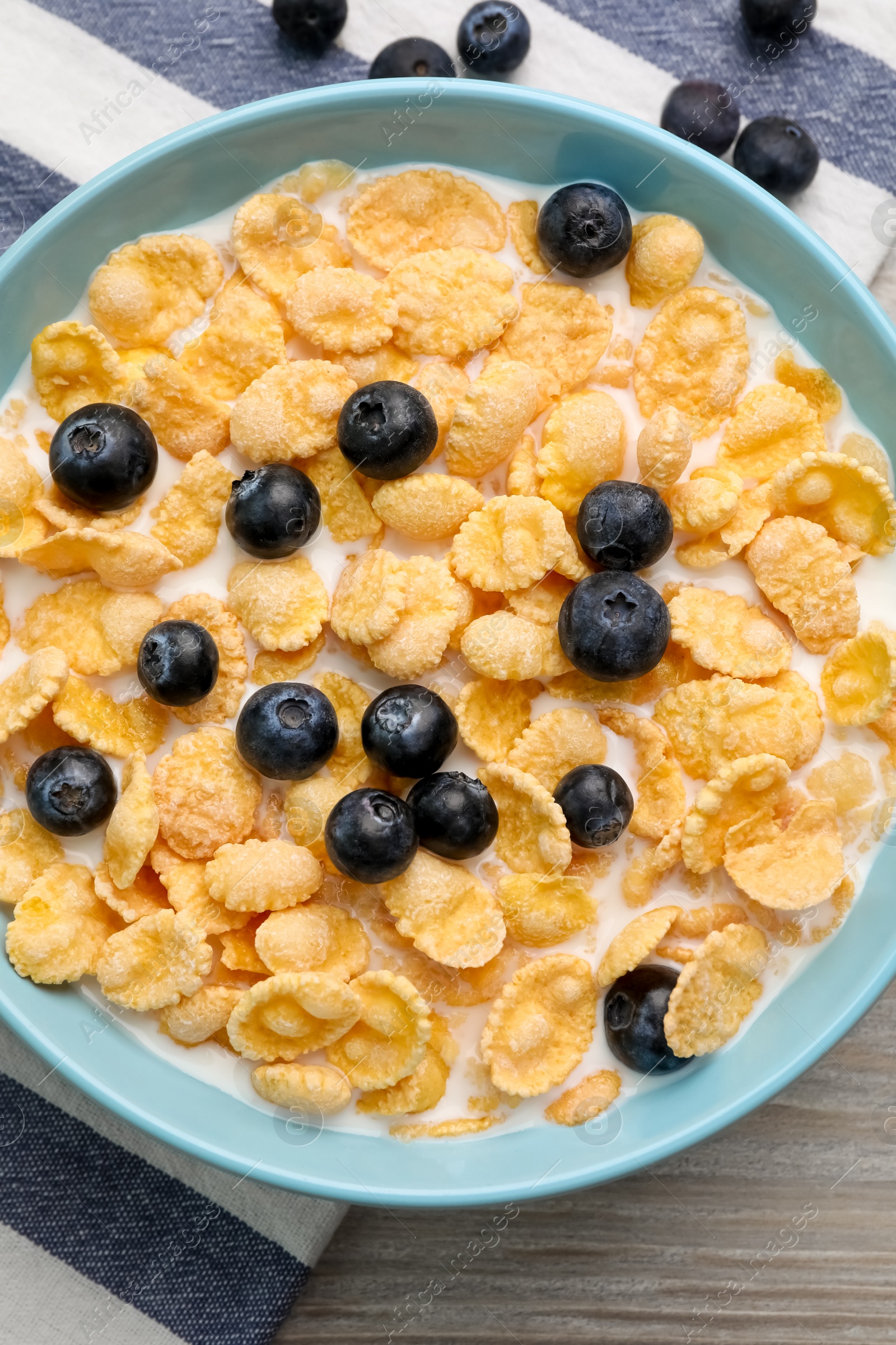 Photo of Bowl of tasty crispy corn flakes with milk and blueberries on wooden table, flat lay