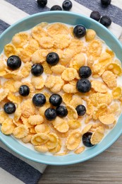 Bowl of tasty crispy corn flakes with milk and blueberries on wooden table, flat lay