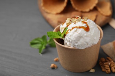 Photo of Tasty ice cream with caramel sauce, mint and nuts in paper cup on grey wooden table, closeup. Space for text