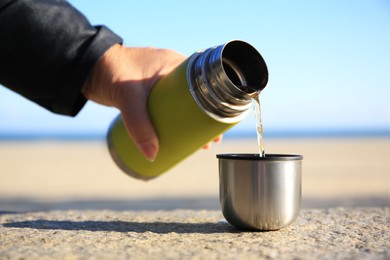 Photo of Woman pouring hot drink from yellow thermos into cap on stone surface outdoors, closeup