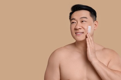 Photo of Handsome man applying cream onto his face on light brown background. Space for text