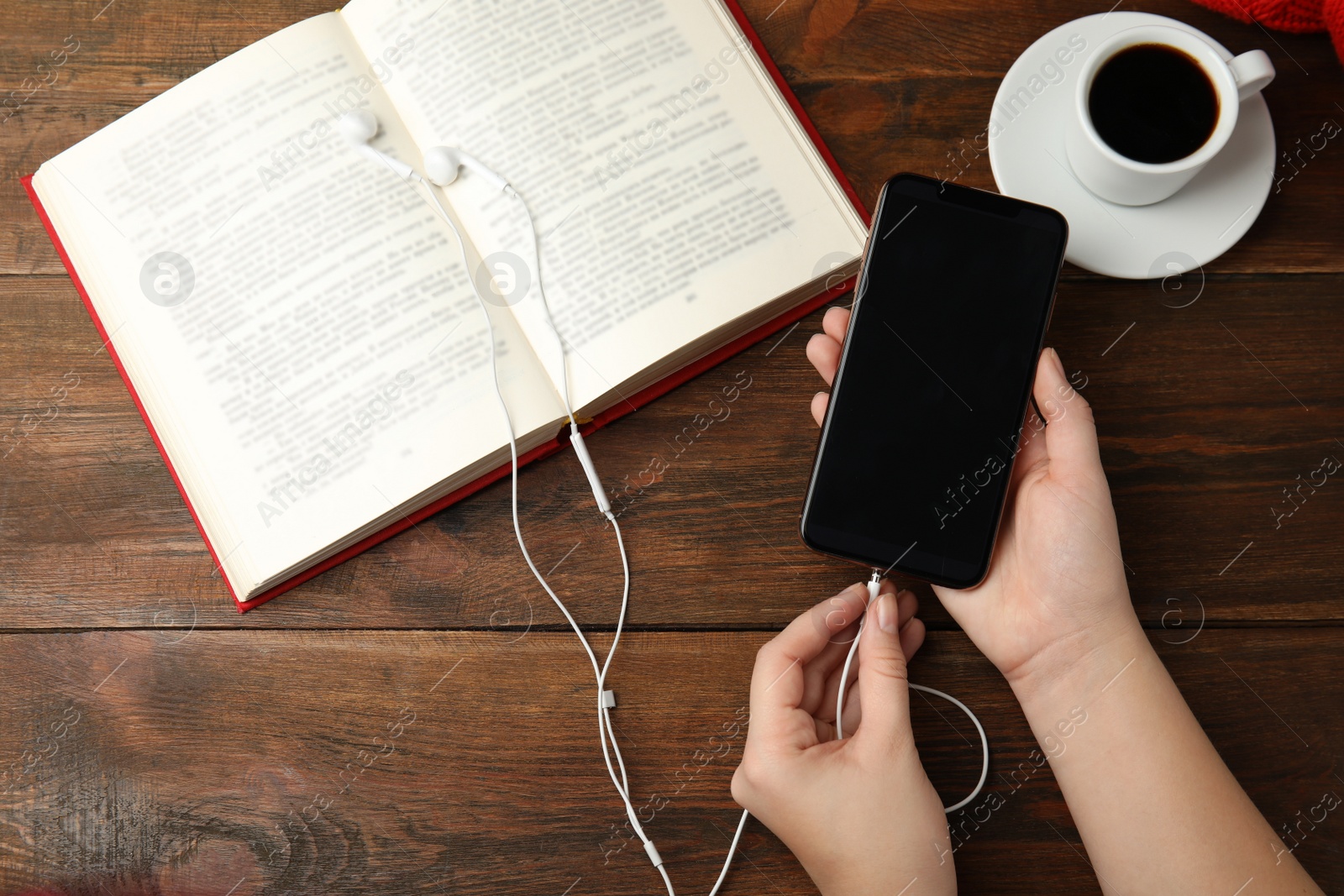 Photo of Woman plugging earphones into mobile phone over wooden table with book, top view