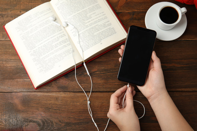 Woman plugging earphones into mobile phone over wooden table with book, top view