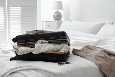 Open suitcase full of clothes, jacket and fashionable accessories on bed in room